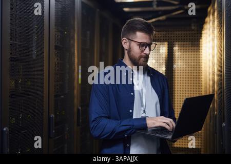 IT professional monitoring server equipment in data center holding laptop for diagnostics and system checks surrounded by racks of servers Stock Photo