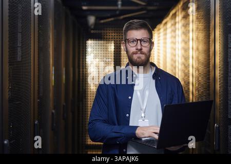Man with beard and glasses holding laptop while inspecting servers in a data center with focused expression seen among rows of server racks. Professional environment with blurred background Stock Photo