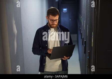 Man with beard and glasses standing in a dark corridor of a data center, working on laptop while inspecting server racks and equipment Stock Photo