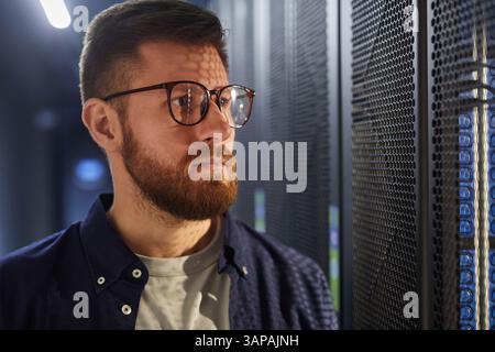 Bearded man standing in a data center, working with servers and equipment, wearing glasses and looking focused. Technology and servers visible in background Stock Photo