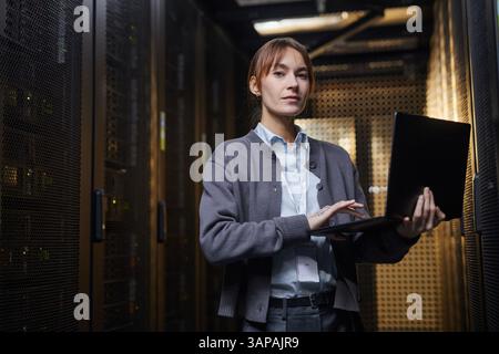 Woman standing in server room holding laptop, concentrating on her work in a high-tech environment filled with racks of servers and equipment Stock Photo