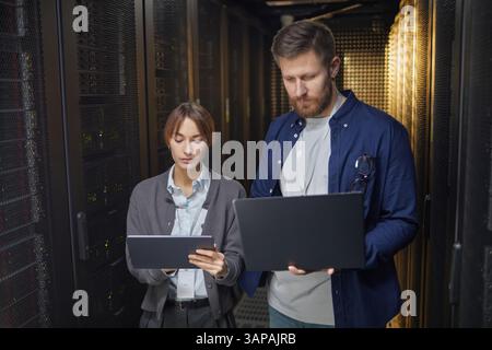 Two IT professionals working together in a data center, using tablets to monitor servers and network equipment creating an efficient and organized environment Stock Photo
