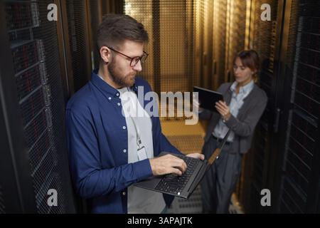 Two IT professionals managing server equipment in modern data center. One person is using laptop while another is using tablet, ensuring all systems are operational Stock Photo