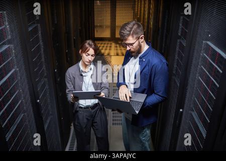 Two IT professionals collaborating in data center while using laptop and tablet, surrounded by server racks. They are deeply engaged in their work, ensuring efficient operations Stock Photo