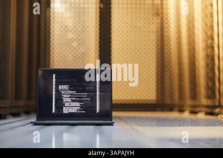 Open laptop displaying lines of code against backdrop of server racks in dim lighting. Scene represents data processing, programming, and technology infrastructure maintenance Stock Photo