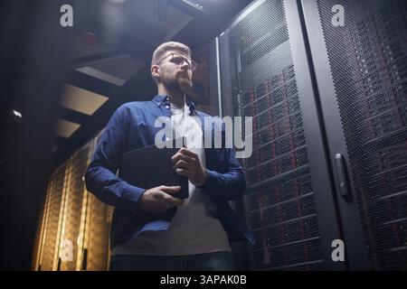 Young male professional inspecting server room holding clipboard wearing glasses and a blue shirt Visible server racks behind showing technological environment Stock Photo