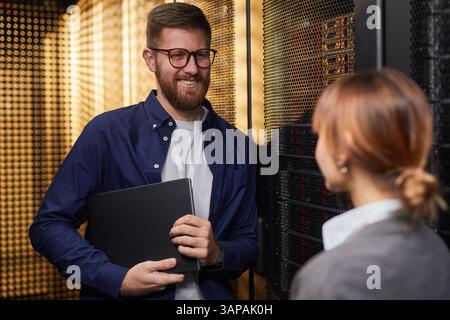 Smiling IT professional holding laptop while talking with colleague in server room surrounded by network equipment working on project Stock Photo