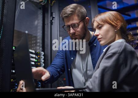Multiracial team working on server maintenance in a data center. Man and woman collaborating while examining server rack and discussing technical details Stock Photo
