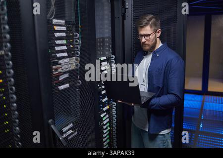 Male IT technician standing in modern server room holding laptop, checking network equipment and ensuring proper functionality. High-tech environment featuring server racks and data cables Stock Photo