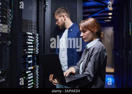 IT professionals in a data center managing server operations while collaborating on tasks with laptops. Focused on server health and data management Stock Photo