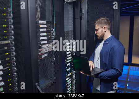 Technician standing in server room checking connections and status of servers using laptop while surrounded by multiple racks of equipment and cables Stock Photo