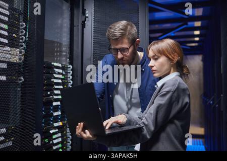 Two IT professionals are examining server connections in a data center, focusing on laptop screen while discussing necessary steps for troubleshooting issues Stock Photo