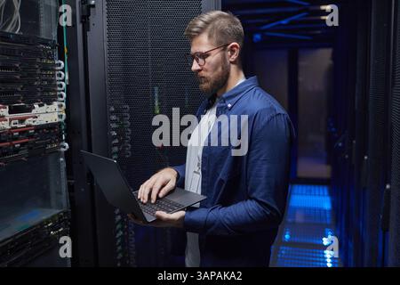 Bearded man in blue shirt engaged in managing servers in a modern data center, holding a laptop while examining server racks for maintenance and monitoring purposes Stock Photo