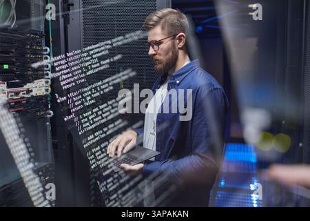 IT professional configuring server settings in modern data center with a laptop. Technical screens displaying code and server racks visible in background Stock Photo