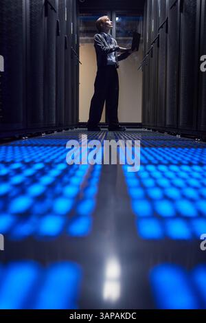 Technician wearing professional attire working on laptop inside server room, surrounded by advanced computer equipment and glowing blue lights on the floor creating a futuristic ambiance Stock Photo