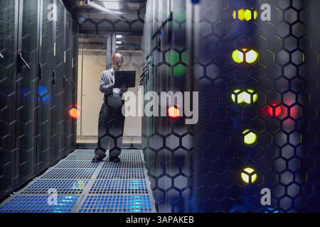 Technician in data center checking system performance on laptop, standing between server racks illuminated by LED indicators Stock Photo