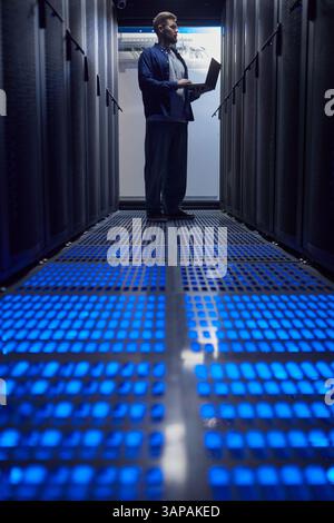 Man holding laptop in a data server room, surrounded by tall server racks, with blue illuminated floor panels creating a futuristic atmosphere Stock Photo
