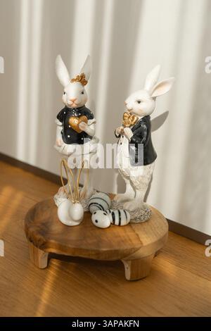 Two ceramic bunny figurines dressed elegantly, holding gold heart ornaments, are displayed on a wooden stand with decorative eggs. The scene captures Stock Photo