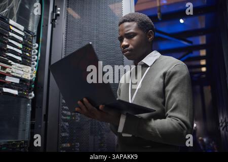 African American technician working with a laptop inside data center server room, analyzing server performance, monitoring systems and infrastructure, ensuring network stability and connectivity Stock Photo