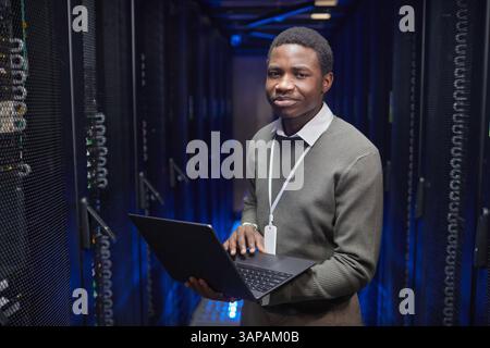 IT professional standing in server room, holding laptop with confident expression. Blue lighting highlights server racks in background, indicating technology environment Stock Photo