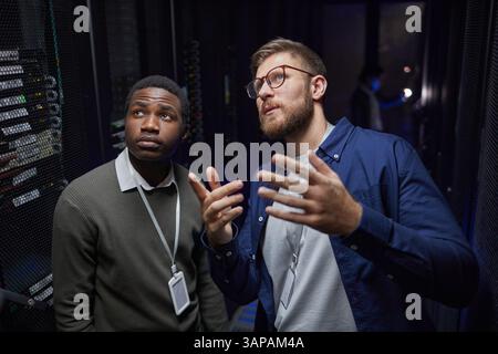 Two technicians in data center, one African American, standing near server racks while discussing maintenance process, one person gesturing with hand, showing attentiveness Stock Photo