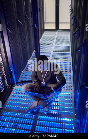 Engineer monitoring server performance while sitting on floor inside data center filled with blue neon lighting. Surrounded by towering server racks, focused on device Stock Photo