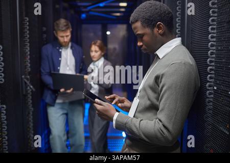 Team of IT professionals working with gadgets in server room analyzing data on devices. Focus on technology and collaboration in high-tech environment Stock Photo