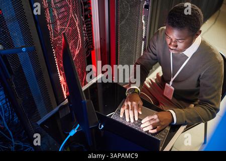 African American IT technician working on laptop in a server room, controlling and maintaining servers, ensuring system functionality, surrounded by hardware infrastructure Stock Photo