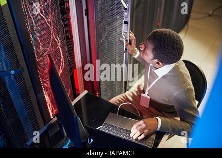 African American man monitoring server room equipment in data center, sitting with laptop, ensuring systems are operational and secure Stock Photo