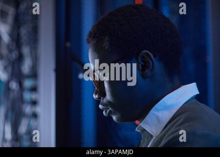 Profile view of a dedicated technician examining computer network in a server room under low light. Intense concentration on technological equipment Stock Photo
