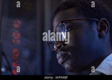 Detail of a person wearing glasses, engaged in data analysis with a backdrop of codes on monitors. The image highlights concentration and technological environment Stock Photo