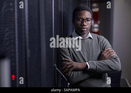 Portrait of young African American man standing in server room, crossing arms against chest and looking straight at camera. Background shows server equipment Stock Photo