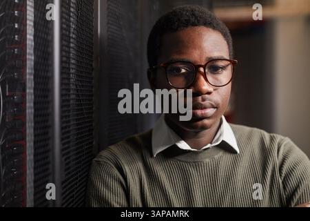 Portrait of young African American man wearing glasses and standing with serious expression. Indoor setting with technology equipment visible Stock Photo