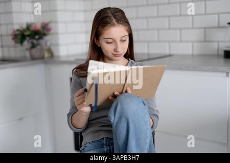 Sweet Teen Girl Reading Notebook Entries While Sitting On A Chair At Home Stock Photo