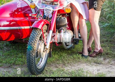 A woman is posing in front of a red motorcycle with a sidecar. The motorcycle is parked on a dirt road, and the woman is wearing red heels. Concept of Stock Photo