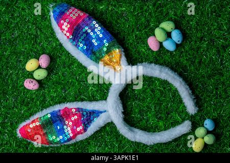 Vibrant bunny ears made of sequins laid on green grass with scattered pastel-colored eggs, perfect for an Easter celebration. Stock Photo