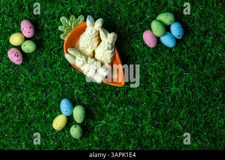 Brightly colored candy bunnies sit in an orange carrot dish, surrounded by pastel chocolate eggs on lush green grass. Stock Photo