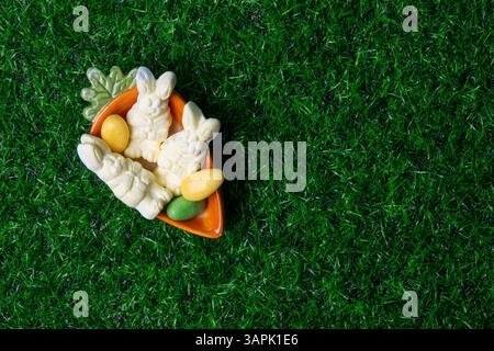 Candy bunnies and vibrant eggs are artfully displayed in a carrot-shaped bowl placed on a grassy surface, evoking a festive mood. Stock Photo