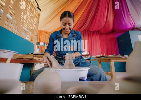 Mexican woman, 24, molds a ceramic piece in her studio wearing a blue apron, shirt, and jeans. Tools and finished pieces surround her. Stock Photo