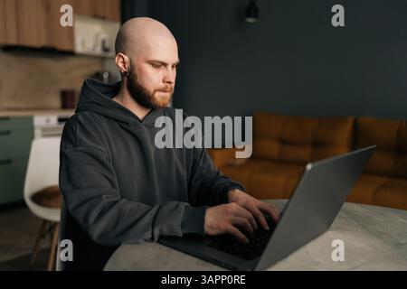 Focused programmer wearing dark hoodie typing code on laptop computer , sitting at table near bright window in contemporary urban apartment Stock Photo