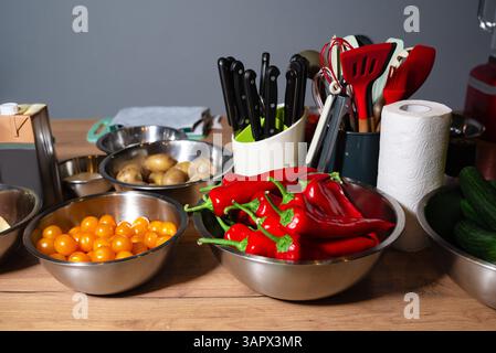 Red peppers, yellow cherry tomatoes, potatoes, and cucumbers in bowls alongside cooking utensils and knives on a kitchen table. Stock Photo