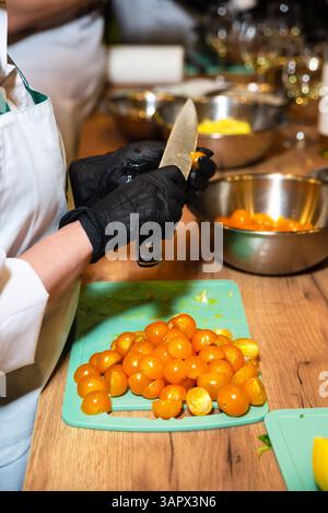 Close-up of a person wearing black gloves cutting fresh yellow cherry tomatoes on a chopping board during food prep. Stock Photo