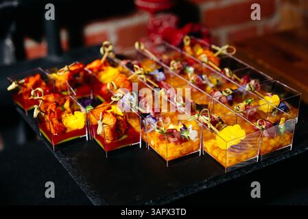 Gourmet appetizer cups filled with mango ceviche and seafood, topped with microgreens, cured meat, and colorful edible flowers on a black tray. Stock Photo