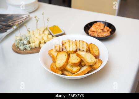 Crispy toasted baguette slices served in a bowl, accompanied by a cheese platter and dipping sauces on a white table. Stock Photo