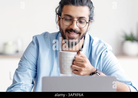 Young Smiling Arab Freelancer Guy Using Laptop And Drinking Coffee At Home Stock Photo