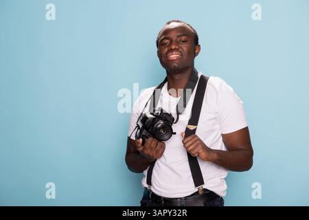 African American man stands confidently, holding a camera in one hand and adjusting his suspenders strap with the other. Portrait of a black male model in casual clothing posing with his DSLR. Stock Photo