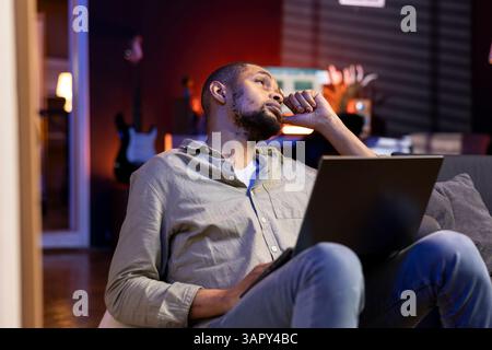 Focused african american guy multitasking on laptop in stylish living space, connected to the internet while managing his freelance work and remote responsibilities. Browsing web laying on sofa. Stock Photo
