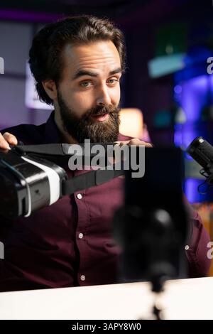 Tech professional reviewing VR glasses in neon lit studio, presenting specifications. Man filming virtual reality headset review for technology fans using cellphone on tripod stabilizer Stock Photo