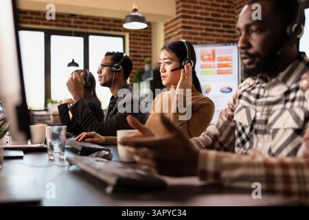 Multiethnic call center agents work at their stations, engaging with remote customers and processing service requests. Computers, coffee cups, and headsets set the scene for a busy office. Stock Photo