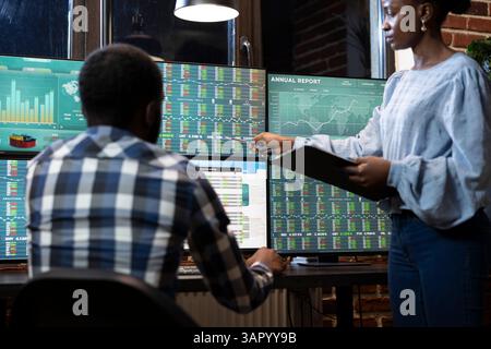 Two traders working late at night in a modern office, collaborating on multiple monitors displaying forex market charts and stock data. African american business partners reviewing stock trading data. Stock Photo